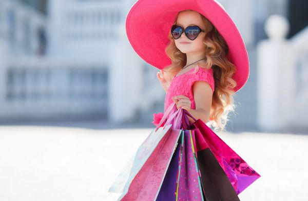 دختر کوچک ناز در کلاه شیک در خرید پرتره یک بچه با کیسه های خرید کودک در لباس عینک آفتابی و کفش در نزدیکی مرکز خرید لذت ببرید خريد كردن دختر روش