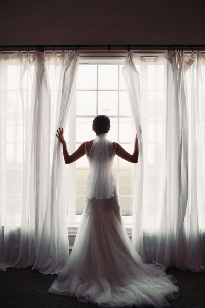 عروس زیبا در لباس عروسی خیره کننده به نظر می رسد پنجره