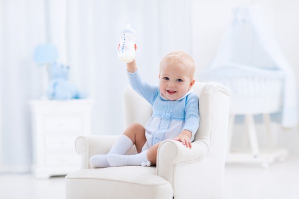 کودک عجیب و غریب در یک کف آبی کفپوش و شیر آشامیدنی از یک بطری در یک مهد کودک آفتابی سفید با صندلی گهوار و اتاق خواب داخلی با کابین نوزاد فرمول نوشیدنی برای نوزاد