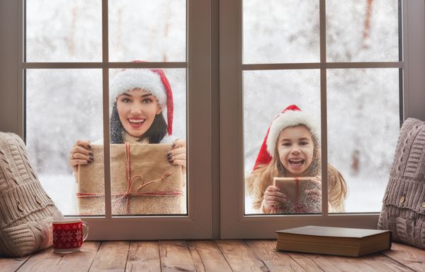 کریسمس مبارک و تعطیلات دختر کوچک ناز و مادر جوان او به دنبال پنجره ایستاده در خارج از منزل در پس زمینه جنگل زمستانی کودک و بزرگسال برگزاری جعبه هدیه حاضر
