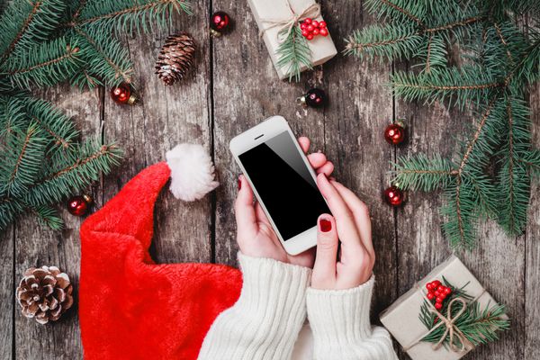 زنان دست نگه داشتن تلفن همراه در پس زمینه چوبی با هدایای کریسمس کلاه سانتا شاخه های Fir کریسمس و ترکیب سال نو مبارک تختخواب نمای بالا
