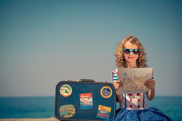 کودک مبارک با چمدان پرنعمت و نقشه شهر در ساحل بچه ها سرگرم کننده در تعطیلات تابستانی پرتره دختر در برابر دریا و پس زمینه آسمان مفهوم سفر و ماجراجویی