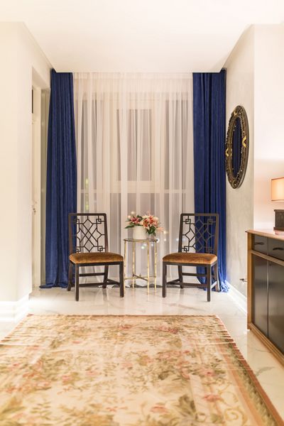 سالن مدرن با دیوارهای سفید و کف کاشی های سبک با فرش با الگوهای گل دو صندلی شیک یک میز کوچک فلزی با گل در یک گلدان آینه فانتزی پایه چوبی با زیر شلواری وجود دارد