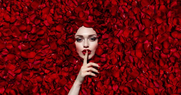 پرتره یک دختر جوان زیبا در گلبرگ گل رز قرمز لب قرمز ناخن های قرمز دختر با حرکاتش دست و پا می زند