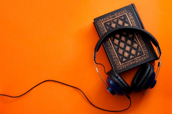 کتاب صوتی پادکست و گوش دادن به مفهوم ادبی با هدفون بر روی یک کتاب پرنعمت در پس زمینه نارنجی رنگی با فضای کپی