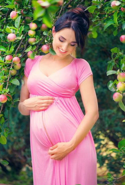 زن باردار شاد کردن شکمش زیبایی باردار پرتره در فضای باز مادر جوان نوازش شکم و لبخند زدن مفهوم بارداری سالم زن باردار در باغ