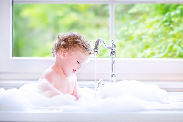 دختر بچه کوچک خنده دار با موهای فرفری مرطوب با استفاده از یک حمام در یک سینک آشپزخانه با مقدار زیاد فوم با قطرات آب و چلپ چلوپ کردن کنار یک پنجره بزرگ با نمایش باغ