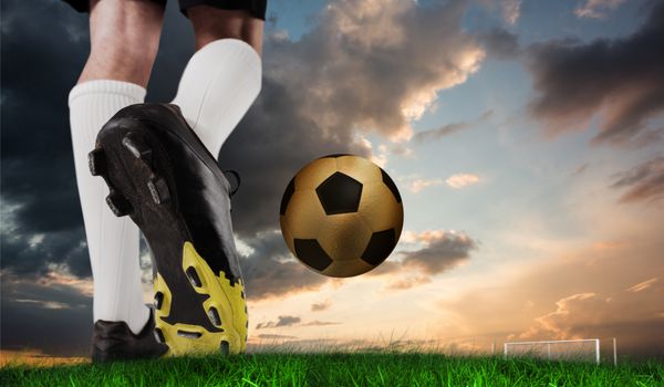 تصویر کامپوزیت از فوتبال بوت لگد زدن توپ طلا در برابر چمن سبز تحت آسمان آبی و نارنجی