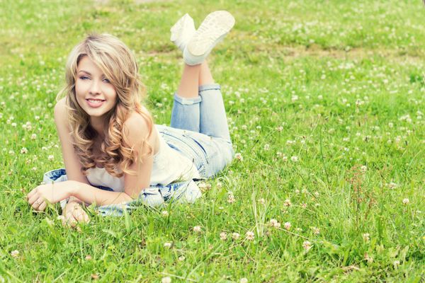 دختر خوش تیپ زیبا که روی چمن دروغ می گوید و لبخند می زند در شلوار جین در روز تابستان آفتابی در باغ