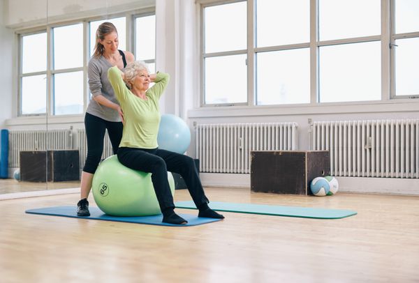 زن ارشد نشسته در یک توپ پیلاتس تمرین در باشگاه سلامت که توسط مربی شخصی اش کمک می کند درمانگر فیزیکی کمک به زن ارشد در تمرین او در ورزشگاه