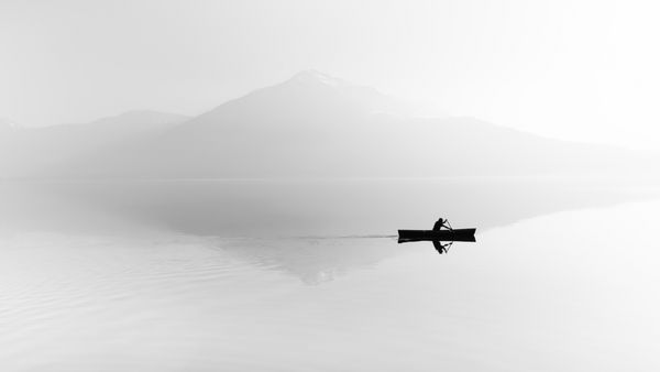 تورم بر روی دریاچه Silhouette کوه ها در پس زمینه مرد در یک قایق با یک دست و پا زدن شناور است سیاه و سفید
