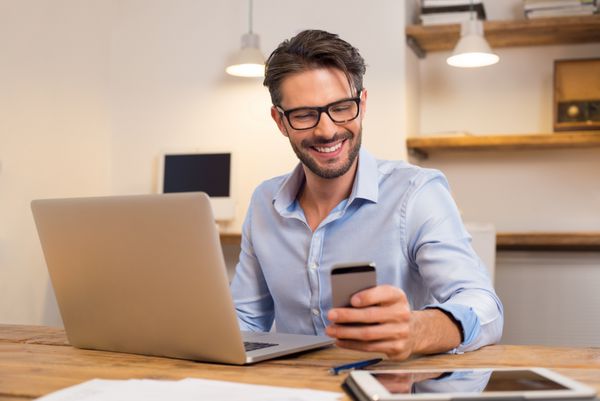 جوان بازرگان خندان گوشی های هوشمند خود را هنگام خواندن پرتره های خندان پیام خواندن مرد کسب و کار با گوشی های هوشمند در دفتر مرد در پشت میز خود را در دفتر کار