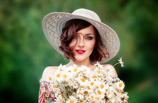 پرتره از مدل زرق و برق دار با یک کلاه و لب قرمز لبخند زدن و نگه داشتن یک دسته گل بزرگ از شاخساره بستن دختر سحر و جادو با بهره گیری از طبیعت در فضای باز Green backgroundArt کار
