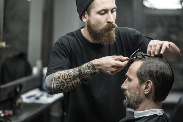 آرایشگر با استفاده از ریشهای بزرگ و اصلاح موهایش بر روی موی مرد در موی سیاه موی سر در آرایشگاه او یک تی شرت سیاه و سفید با کلاه سیاه پوشانده است افقی