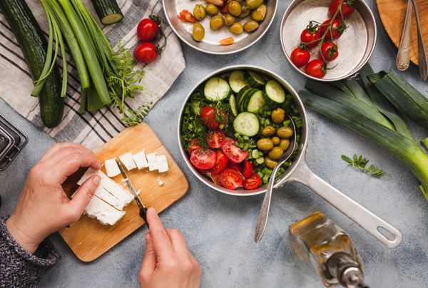 پیاز سبز بهاره خیار و گوجه فرنگی قرمز سرآشپز پنیر فتا را برای سالاد یونانیان بریزید سبزیجات تازه بر روی میز آبی سنگ مرمر نمای بالا