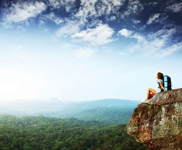 زن جوان با کوله پشتی نشسته روی صخره و به دنبال یک آسمان