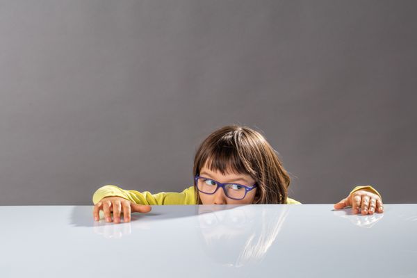 مفهوم آموزش و پرورش با کودک نابینا با عینک پنهان کردن نیمی از صورت زیر جدول نگاه کردن به طرف برای ایده برای فرار peekaboo با استفاده از تخیل سرگرم کننده پس زمینه خاکستری