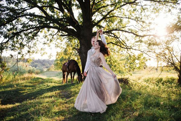 عروس در یک لباس صورتی ظریف با پاکت بر روی یک اسب قهوه ای داماد در یک کت و شلوار سبک با کراوات کمان است عروسی در جنگل در غروب آفتاب پیاده روی در طبیعت در نزدیکی آب