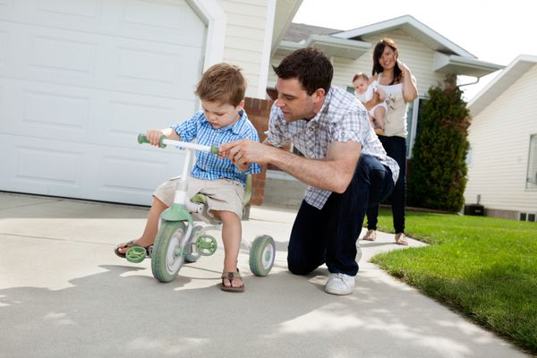 پدر پسر پسر خود را به سوار شدن با سه چرخه در حالی که همسر ایستاده در پس زمینه