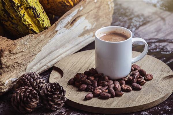 شکلات داغ در فنجان سفید روی میز چوبی در کنار دانه های کاکائو