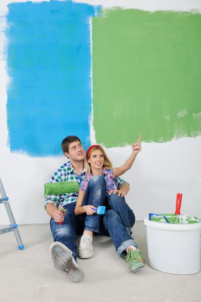 زن و شوهر جوان پس از رنگ آمیزی دیوار سفید با رنگ سبز و آبی در خانه جدیدشان استراحت می کنند