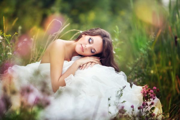 عروس پری فوق العاده در یک لباس عروسی در میان گلهایی با چشمان بسته در چمن سبز قرار دارد