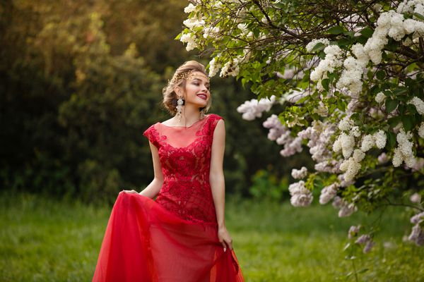 زن زرق و برق دار زیبا با لباس قرمز قرمز در باغ گل یاس شکوفه پرتره رشد در نور غروب خورشید دختر جوان جوان قفقازی در باغ قدم می زند و با لباس خود بازی می کند زمان بهار ملکه جوان