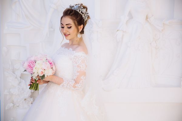 دختری با لباس عروسی سفید با توری که در یک اتاق سفید برفی با مجسمه ها قرار می گیرد دسته گلهای کوچک خود را تحسین می کند
