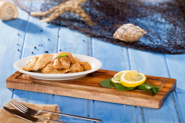 غذاهای دریایی خوشمزه دریایی باس در بشقاب سفید با arugula شوید و روغن زیتون روی تخته برش بامبو که با شبکه ماهیگیری پوسته چاقو چنگال و دستمال در زمینه سفره چوبی آبی روستایی سرو می شود