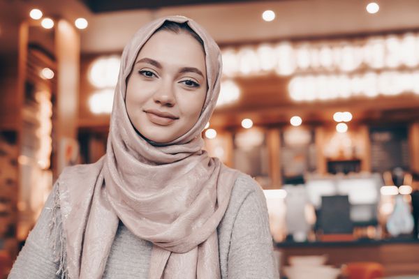 دختر مسلمان زیبا در حجاب که لبخند می زند منتظر غذای خود در یک رستوران است