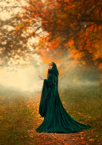 غریبه مرموز شبح در یک مسیر در جنگل چرخانده در یک روپوش سبز زمرد با یک کلاه و آستین های پهن در جنگل پاییزی مه آلود نارنجی قرمز ایستاده یک جادوگر با موهای قرمز آتشین