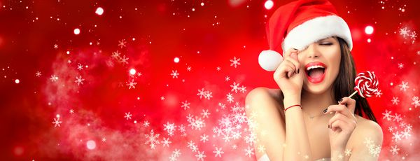 زن کریسمس دختر مدل زیبایی در کلاه بابا نوئل با لب های قرمز و آب نبات شیرین ابریشمی در دست خود شادی ابراز تعجب پرتره نزدیک روی پس زمینه گسترده برف زمستانی قرمز با copyspace