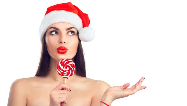 زن کریسمس دختر مدل زیبایی در کلاه بابا نوئل با لب های قرمز و آب نبات آب نبات چوبی lollipop با اشاره ارائه محصول حراجی ابراز تعجب پرتره closeup که در پس زمینه سفید جدا شده است