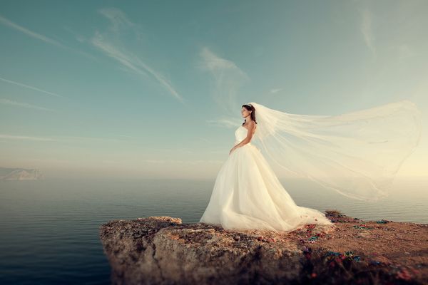 عروس زیبا با لباس عروسی پر زرق و برق بر روی صخره ای بالاتر از دریا ایستاده است