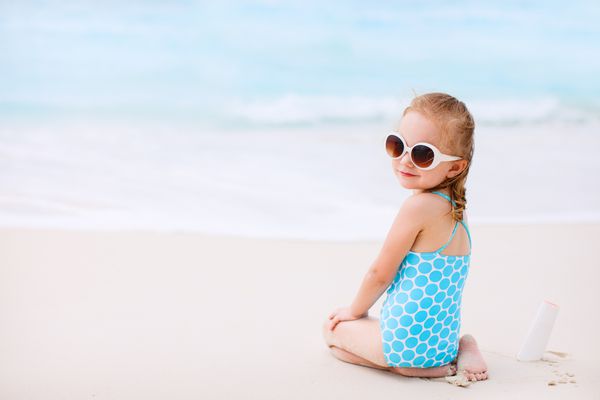 دخترک با بطری کرم خورشید که در ساحل گرمسیری نشسته است