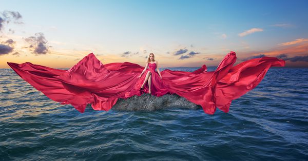 زن جوان در لباس مجلل و بلند لوکس و قرمز قرمز که در دریا نشسته است