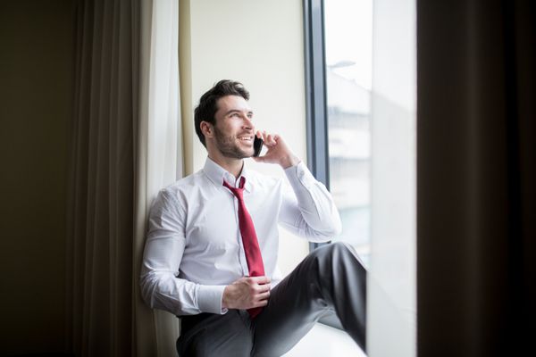 مردی با لباس شخصی که در کنار پنجره نشسته و با تلفن همراه صحبت می کند