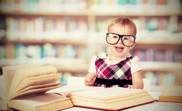 دختر بچه خنده دار در عینک خواندن کتاب در یک کتابخانه
