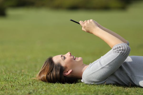 زنی که از یک تلفن هوشمند در حال استراحت روی چمن در یک پارک با پیش زمینه روشن استفاده می کند