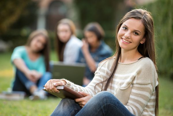 پرتره یك دختر خوشگل و خندان دانشگاهی در حال برگزاری كتاب با دانش آموزان تاری است كه در پارک نشسته اند او به دوربین نگاه می کند