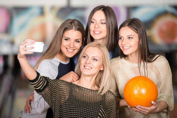گروهی از دوستان جوان زن در یک باشگاه بولینگ در حال گرفتن عکس از خود با تلفن هستند
