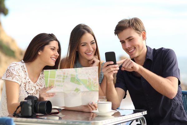 گروهی از دوستان توریستی جوان که مشغول مشاوره نقشه GPS در یک تلفن هوشمند در یک رستوران با ساحل در پس زمینه هستند