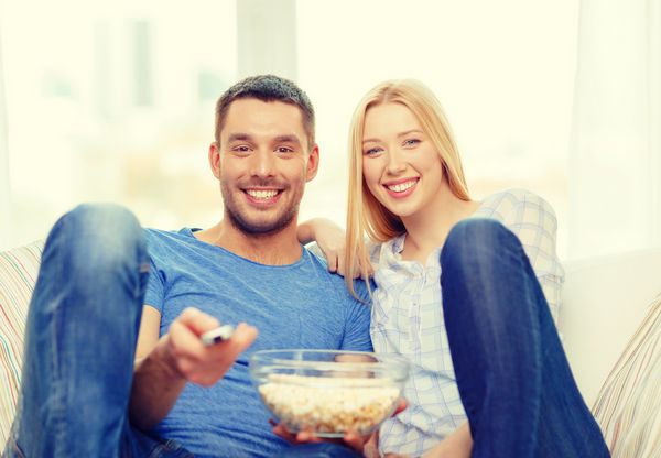 مفهوم غذا عشق خانواده و خوشبختی زن و شوهر با لبخند با پاپ کورن در حال تماشای فیلم در خانه هستند