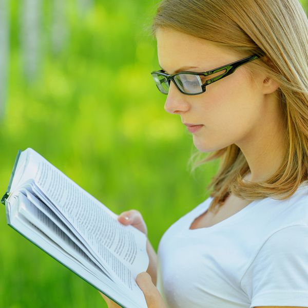 پرتره کتاب زن جوان جذاب لباس های سفید و عینک های عینکی ایستاده در پارک سبز تابستانی