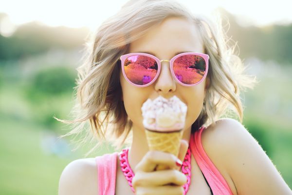 دختر جوان hipster که با خوردن بستنی خوشمزه در هوای تابستان در عینک آفتابی سرگرم کننده و خلق و خوی خوب به نظر می رسد به دوربین و لبخند می زند