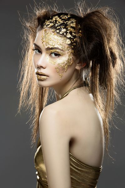 زن جوان و جوان زیبا در لباس با آرایش فویل طلایی هنر روی صورت و لب ها بیش از پیش زمینه خاکستری