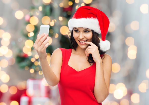 مردم تعطیلات کریسمس و مفهوم فناوری زن زیبا با کلاه سانتا قرمز گرفتن عکس سلفی توسط تلفن های هوشمند بر روی زمینه چراغ