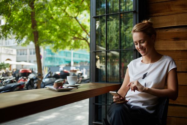 زن جذاب با لبخند زیبا هنگام خواندن خبرهای خوب از طریق تلفن همراه هنگام استراحت در کافی شاپ زن قفقازی که هنگام استراحت در کافه در زمان استراحت در کافه در حال تماشای عکس خود از طریق تلفن همراه است