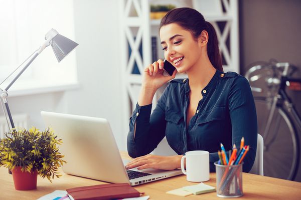 اخبار تجاری خوب را به اشتراک بگذارید زن جوان جذاب که هنگام صحبت در تلفن همراه صحبت می کند و لبخند می زند در محل کار خود در دفتر است و به لپ تاپ نگاه می کند