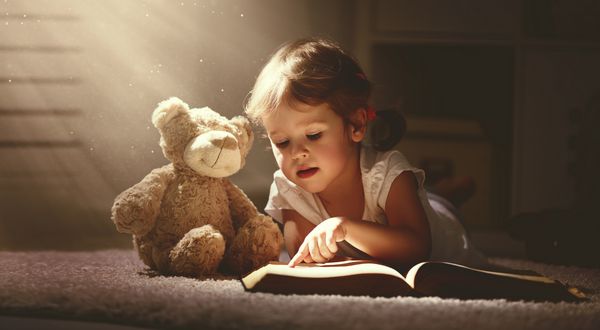 دختر بچه کودک در حال خواندن کتاب جادویی در خانه تاریک با خرس عروسکی اسباب بازی است